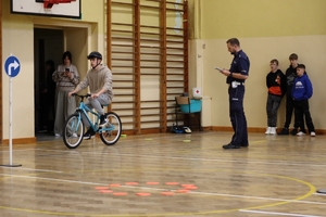 Zdjęcie przedstawia ucznia podczas pokonywania przeszkód na rowerze oraz służby mundurowe podczas sędziowania